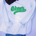 A white crewneck sweatshirt featuring a green glittery 'Cheer' script graphic on the chest, styled with a black ribbed knit beanie and blue jeans against a light lavender background.