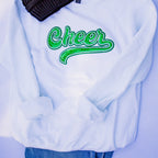 A white crewneck sweatshirt featuring a green glittery 'Cheer' script graphic on the chest, styled with a black ribbed knit beanie and blue jeans against a light lavender background.