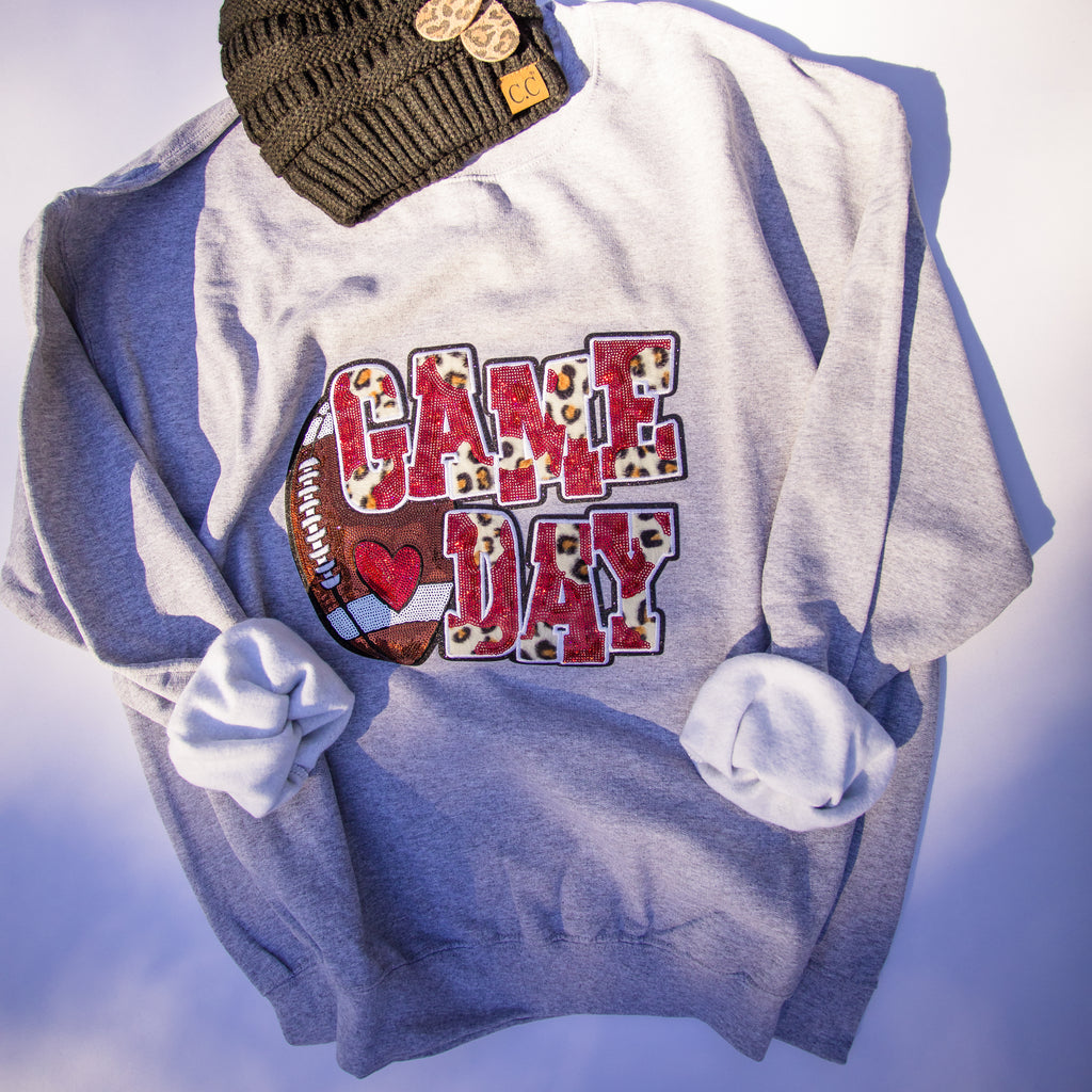 A light gray crewneck sweatshirt with a 'GAME DAY' football graphic in leopard print and red, styled with a dark olive green knit beanie on a white background.