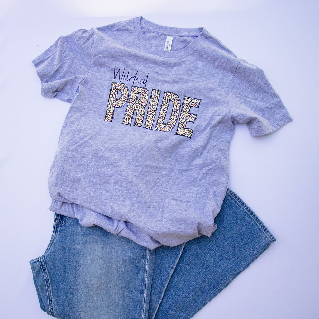 A heather gray short-sleeve t-shirt with 'Wildcat PRIDE' printed across the chest in glittery lettering, styled flat-lay with blue jeans on a white background.