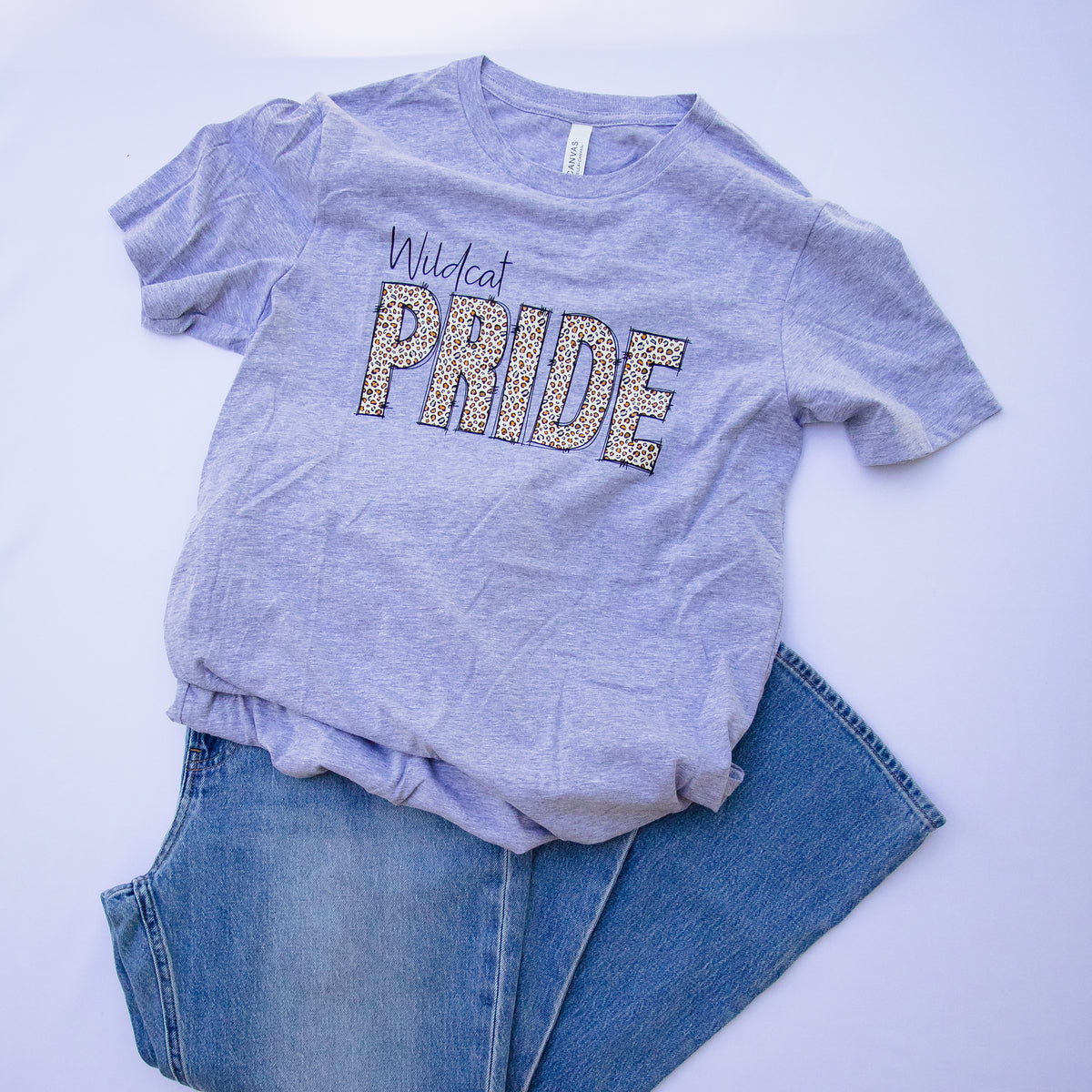 A heather gray short-sleeve t-shirt with 'Wildcat PRIDE' printed across the chest in glittery lettering, styled flat-lay with blue jeans on a white background.