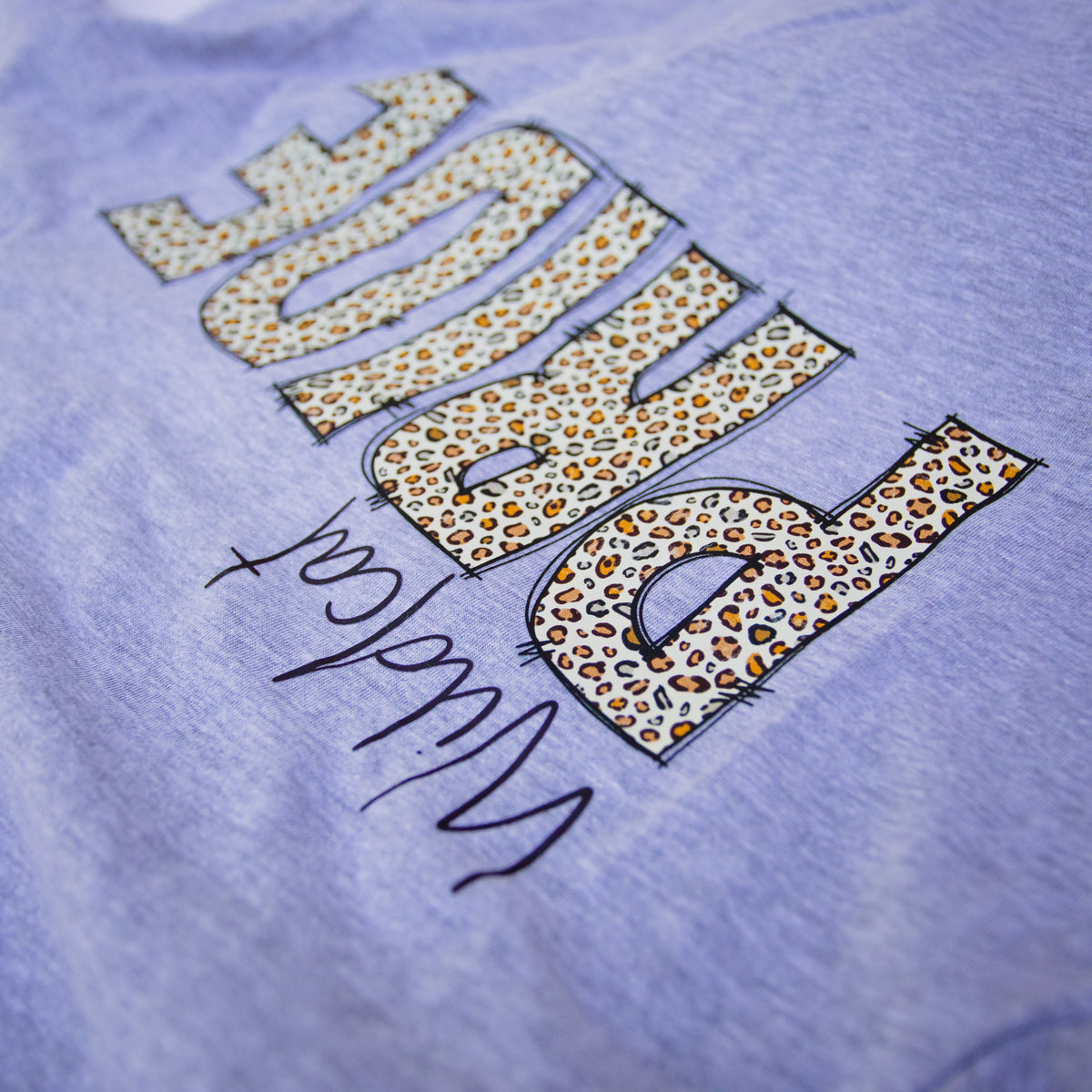 A close-up of a heather lavender-purple t-shirt featuring a graphic print of the word 'PROUD' in leopard print letters with black hand-drawn style outlines, and 'Wildcat' written in black script below.
