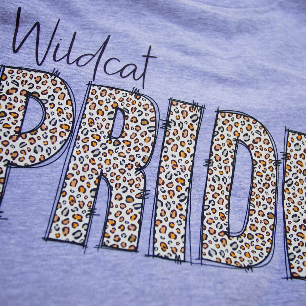 Close-up of a light purple heathered t-shirt featuring a graphic print with the word 'Wildcat' in black script and the letter 'PRIDE' filled with an orange, black, and white leopard print pattern.