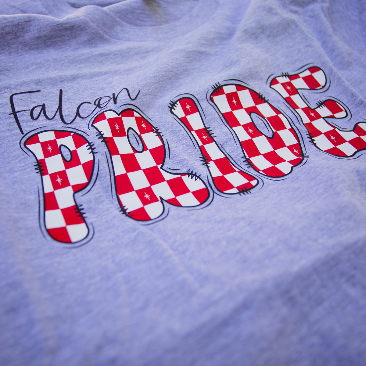 A close-up of a light lavender heather gray t-shirt featuring a graphic print that reads 'Falcon PRIDE'. The word 'PRIDE' is styled in large, bold letters filled with a red and white checkered pattern resembling the Quebec flag, with black stitched details and black outlines.