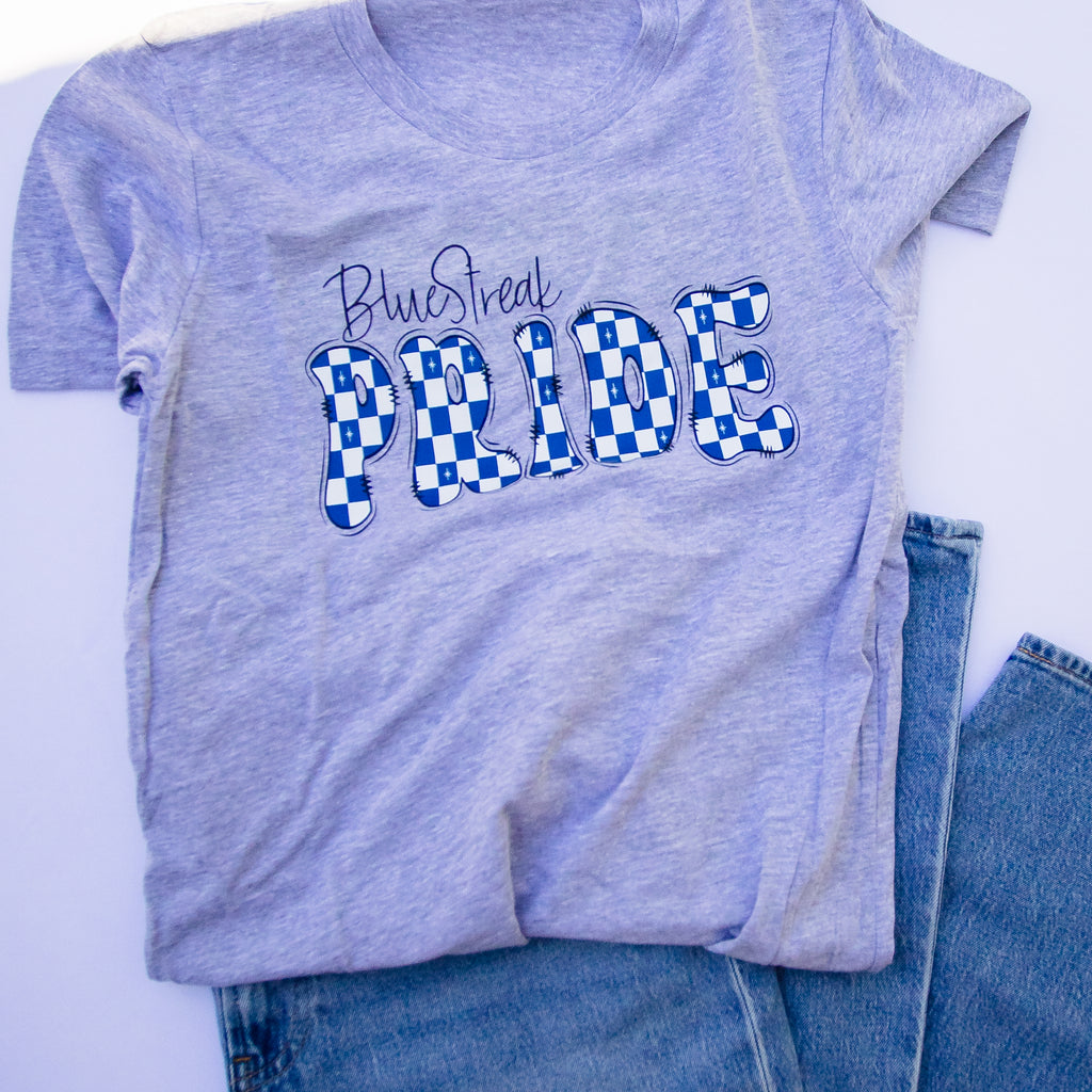 A heather light blue t-shirt with a 'Blue Streak PRIDE' graphic in blue and white checkered pattern, styled flat-lay with blue jeans on a white background.