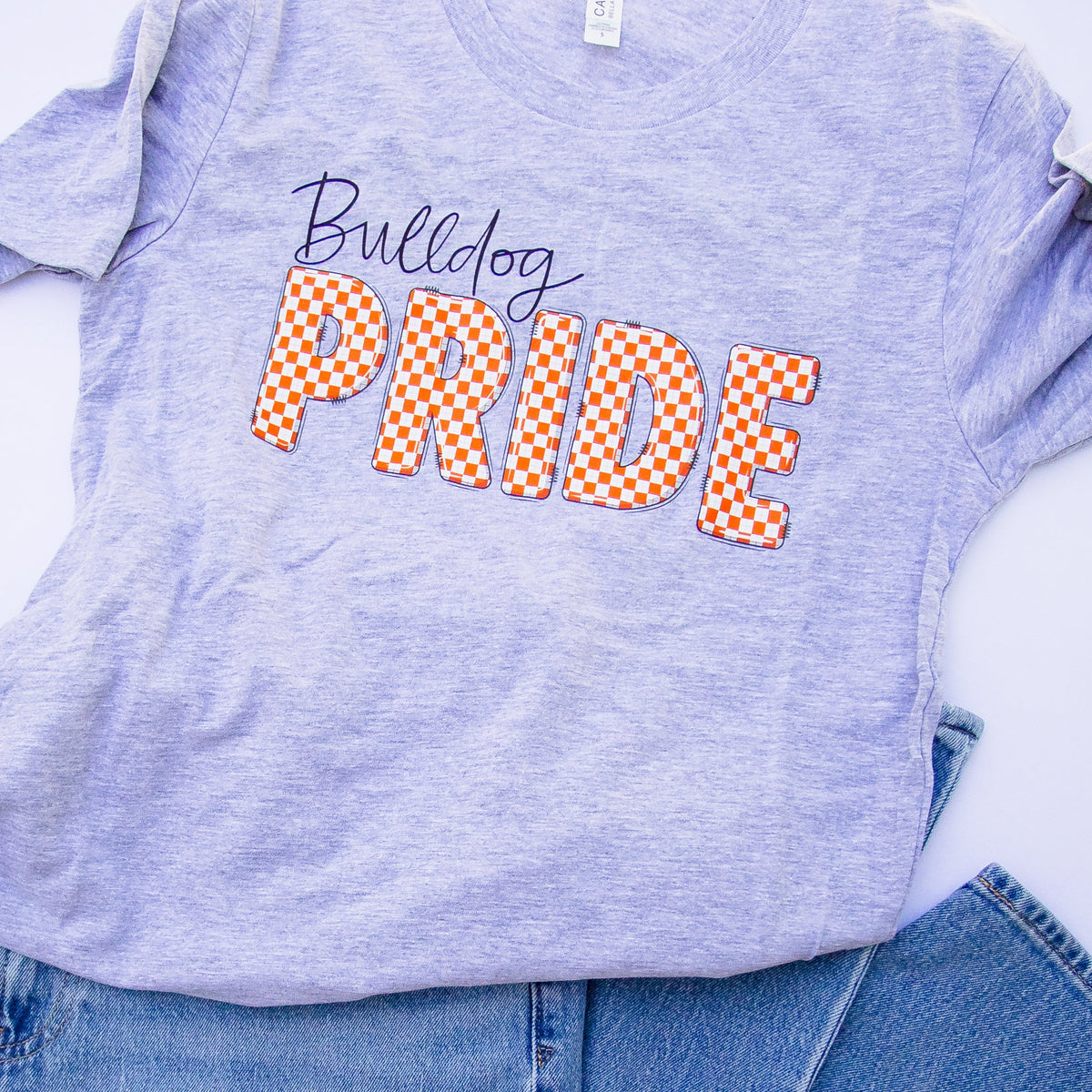 A light heather purple t-shirt with 'Bulldog PRIDE' printed on the front, styled flat-lay with blue jeans on a white background.