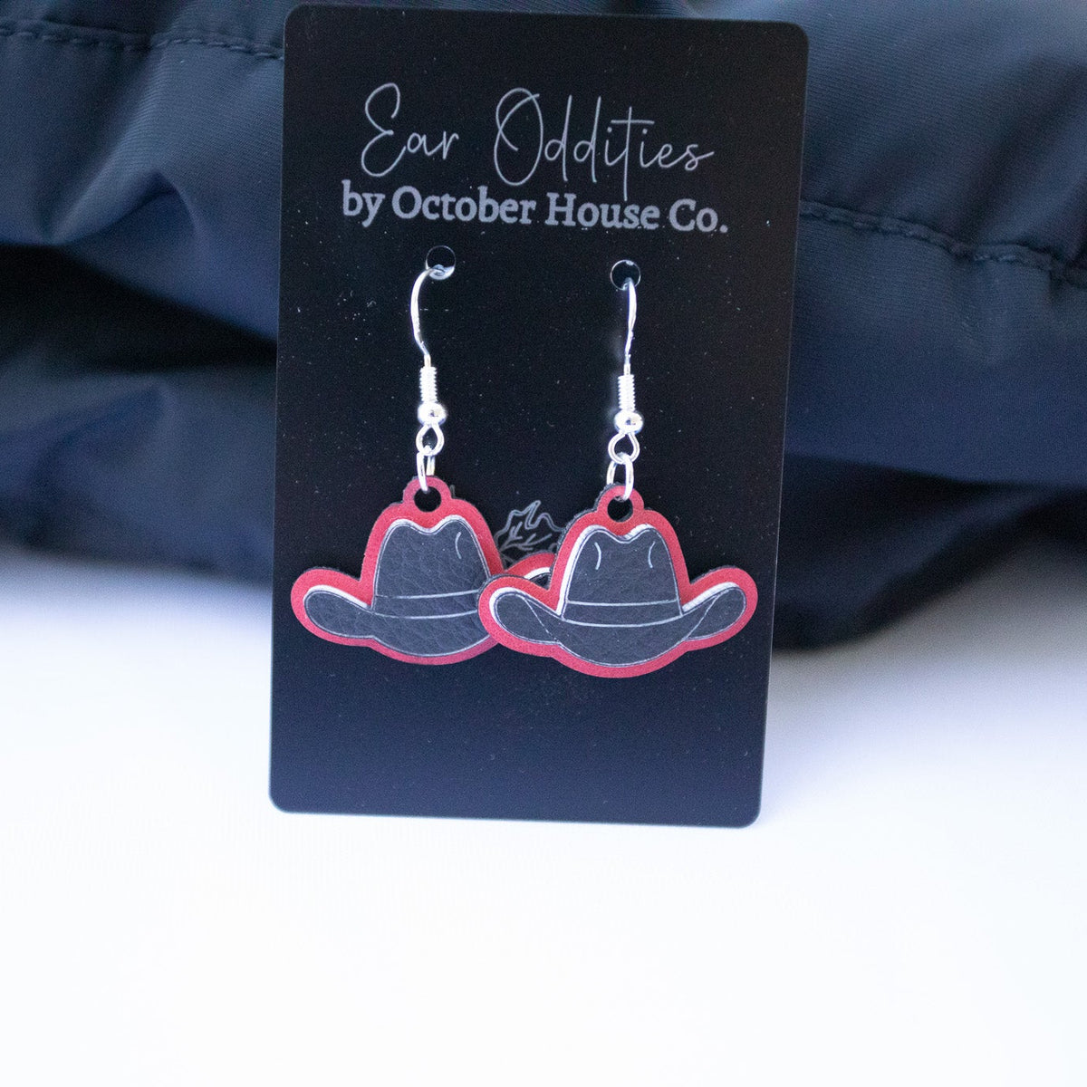 A pair of cowboy hat-shaped dangling earrings with silver hooks and red outlines, displayed on a black card labeled 'Ear Oddities by October House Co.' against a dark fabric background.
