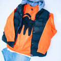 A styled flat-lay of a children's layered outfit featuring an orange sweatshirt with a black cat graphic, a navy blue puffer vest, a gray hooded sweatshirt, and light blue jeans on a white background.