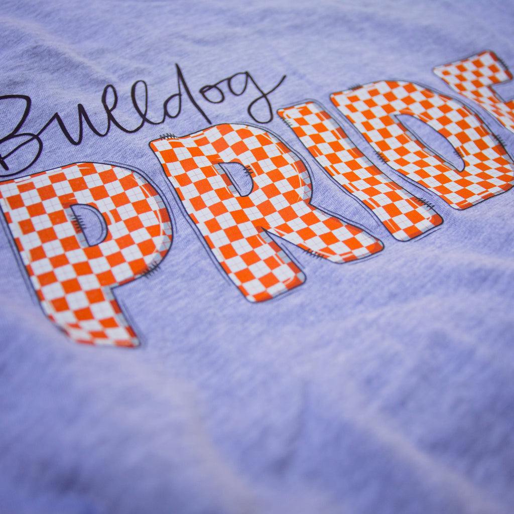 Close-up of a light lavender-purple t-shirt featuring a large orange and white checkered 'PRIDE' graphic with a black script 'Bulldog' above it.