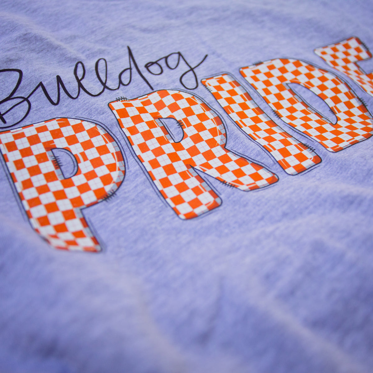Close-up of a light lavender-purple t-shirt featuring a large orange and white checkered 'PRIDE' graphic with a black script 'Bulldog' above it.