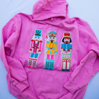 A bright pink hoodie laid flat on a light blue background, featuring three large embroidered nutcracker figures across the chest in pink, yellow, and blue.