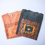 Two folded graphic T-shirts on a light background. The front shirt is charcoal gray with an orange and yellow 'Browns' football graphic.