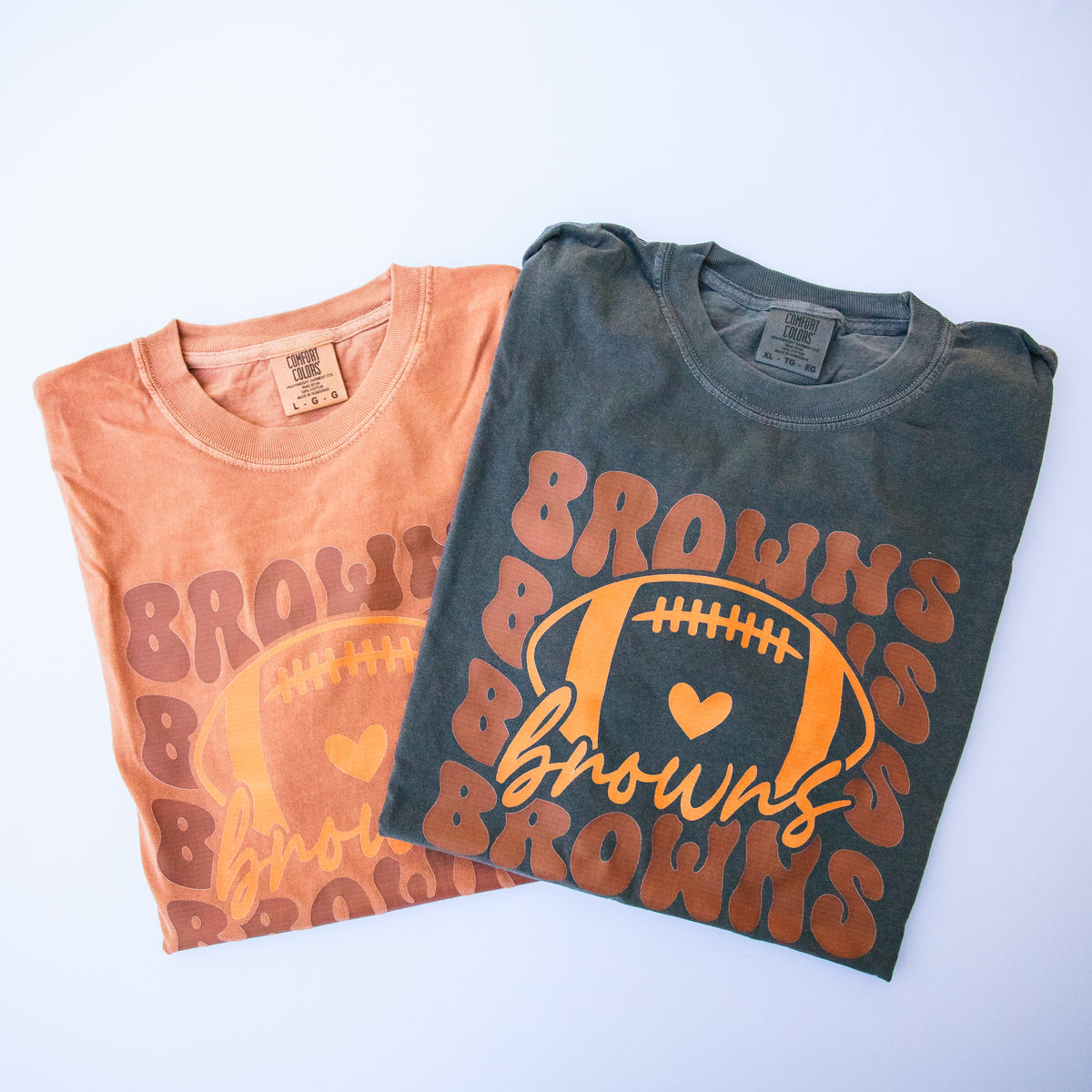 Two folded graphic T-shirts on a light background. The front shirt is charcoal gray with an orange and yellow 'Browns' football graphic.