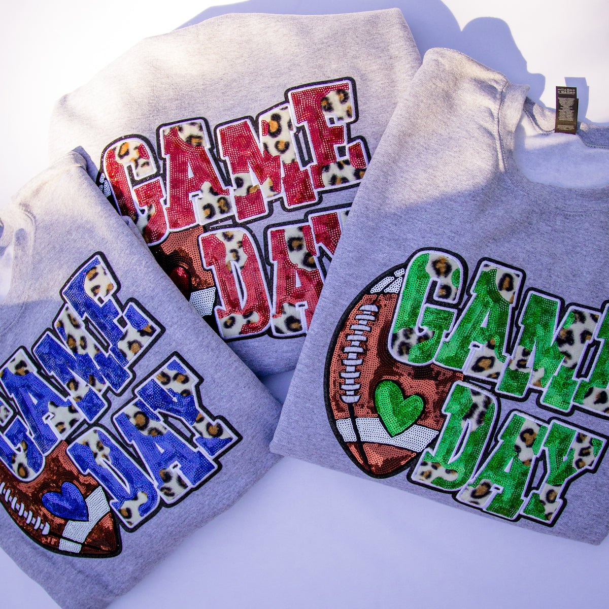 Three light gray sweatshirts arranged together on a white background, each featuring a large 'GAME DAY' graphic with leopard-print lettering and football-themed embellishments in different color combinations.