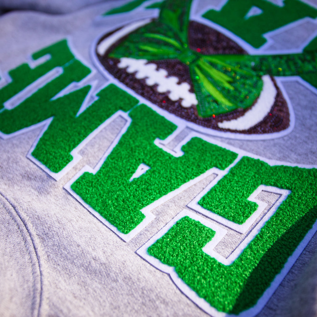 Close-up of a heather gray sweatshirt featuring a green lettering design with a white outline, alongside a green and brown football graphic with white laces.