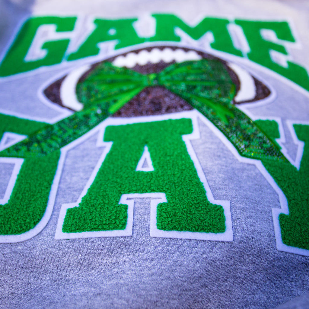Close-up of a gray athletic-style graphic on a garment featuring large green letters outlined in white and a football with a green bow design.