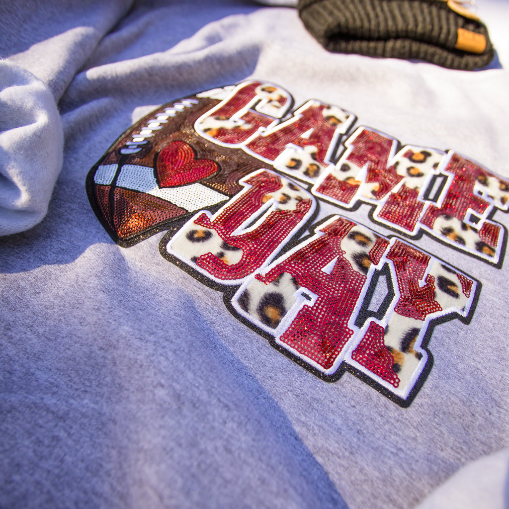 A close-up of a light gray heather t-shirt featuring a large 'GAME DAY' graphic. The letters are filled with a leopard print pattern and outlined in black, with a football graphic on the left containing a red heart and white stripes.