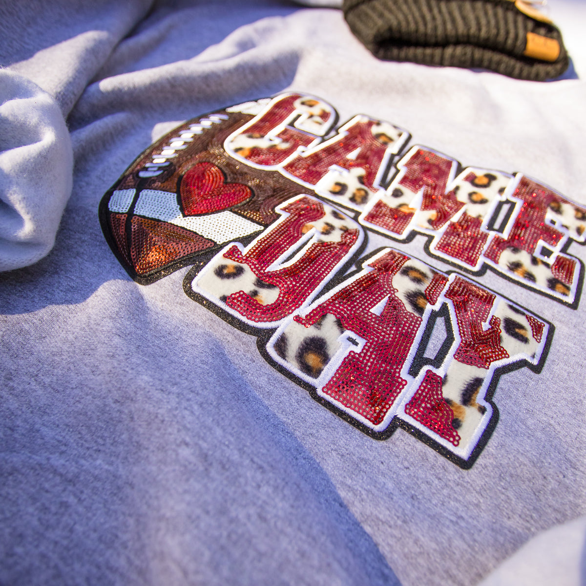 A close-up of a light gray heather t-shirt featuring a large 'GAME DAY' graphic. The letters are filled with a leopard print pattern and outlined in black, with a football graphic on the left containing a red heart and white stripes.