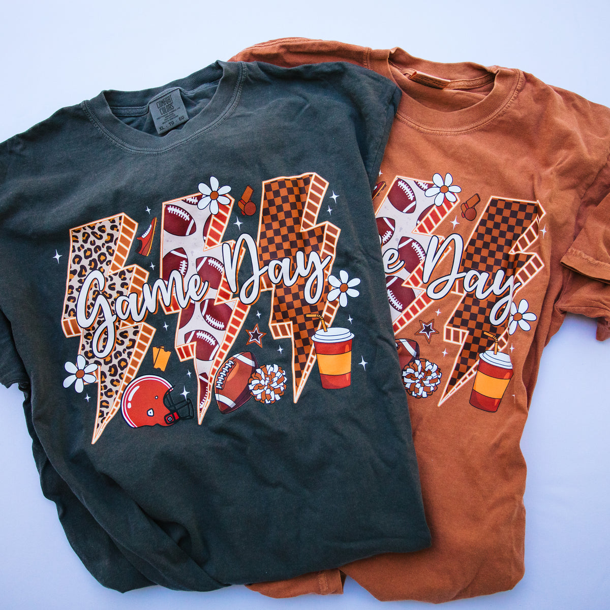 Two graphic t-shirts laid flat on a light blue background. The shirt on the left is charcoal gray, and the shirt on the right is rust brown.
