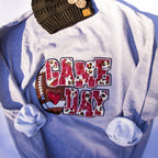 A heather gray sweatshirt with rolled sleeves, featuring a 'GAME DAY' graphic on the front with a football and leopard-print lettering. A dark olive green knit beanie with a small tan 'CC' patch is tucked at the top.