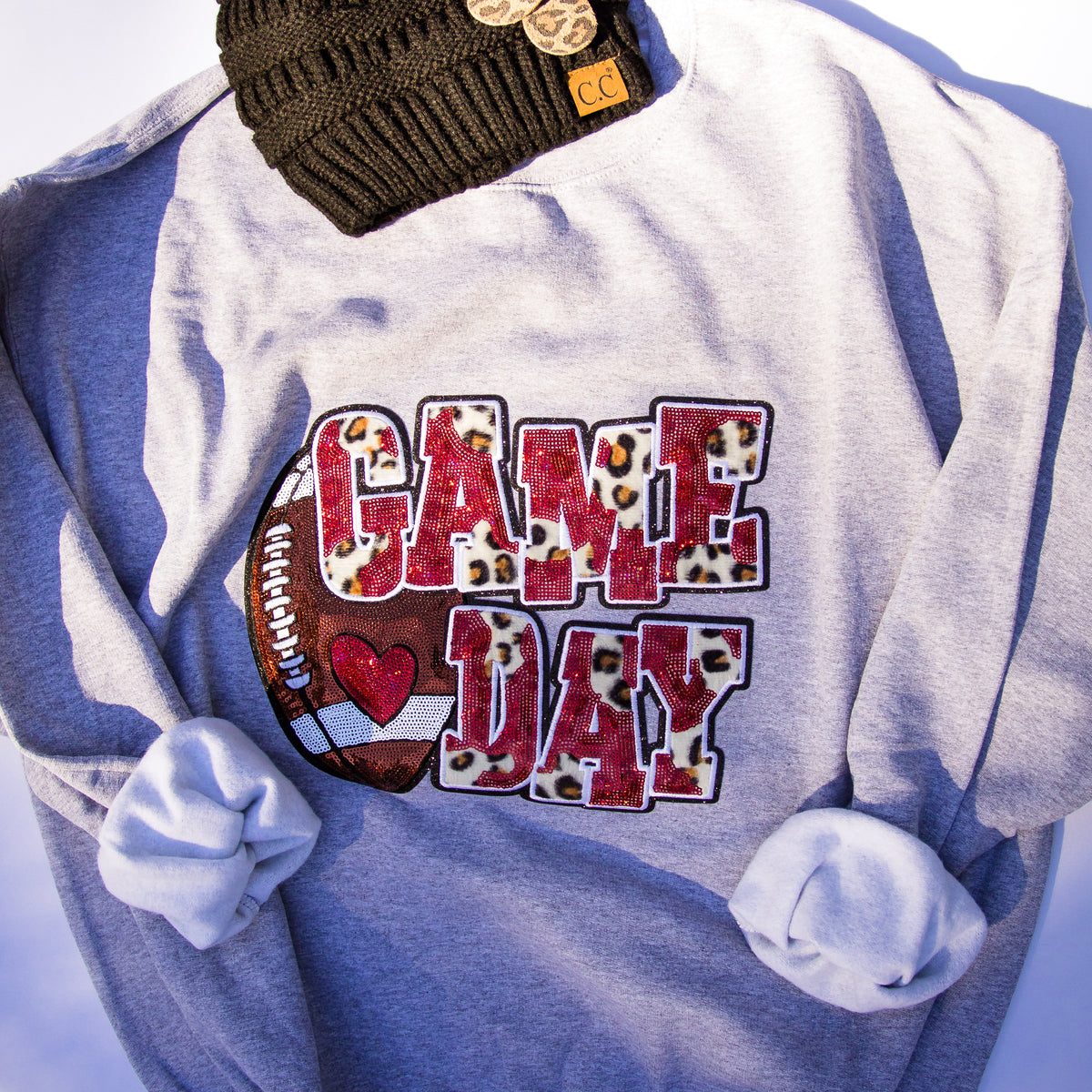 A heather gray sweatshirt with rolled sleeves, featuring a 'GAME DAY' graphic on the front with a football and leopard-print lettering. A dark olive green knit beanie with a small tan 'CC' patch is tucked at the top.