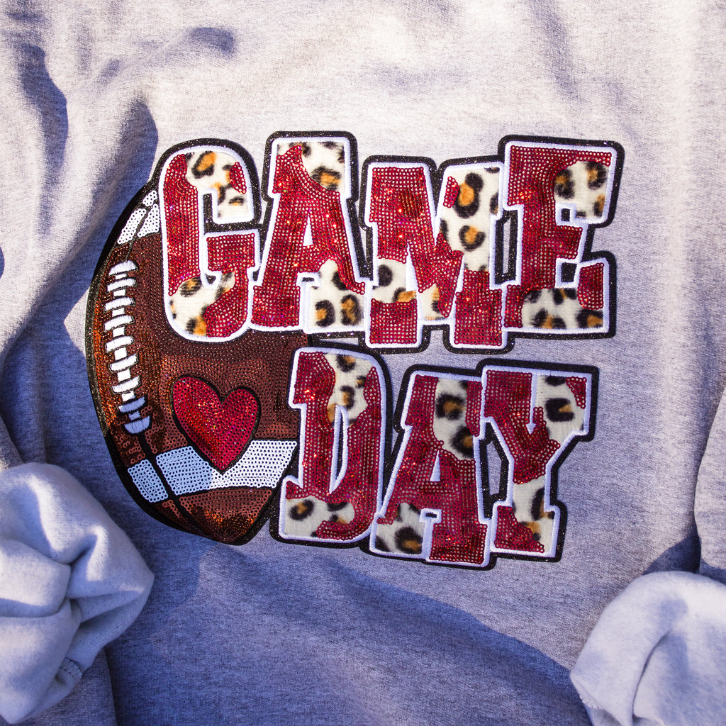 A close-up of a heather gray t-shirt featuring a 'GAME DAY' graphic. The text 'GAME DAY' is styled with leopard print fillings and red glittery lettering, accompanied by a brown football and a red heart graphic.