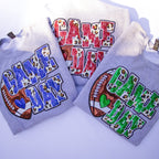Three folded light gray hoodies arranged on a white background, each with a large 'GAME DAY' graphic. One hoodie has blue letters with leopard-print fill and a football motif; another has red letters with leopard-print fill and a football motif; the third has green letters with leopard-print fill and a football motif.