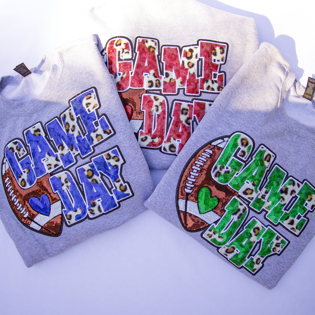 Three folded light gray hoodies arranged on a white background, each with a large 'GAME DAY' graphic. One hoodie has blue letters with leopard-print fill and a football motif; another has red letters with leopard-print fill and a football motif; the third has green letters with leopard-print fill and a football motif.