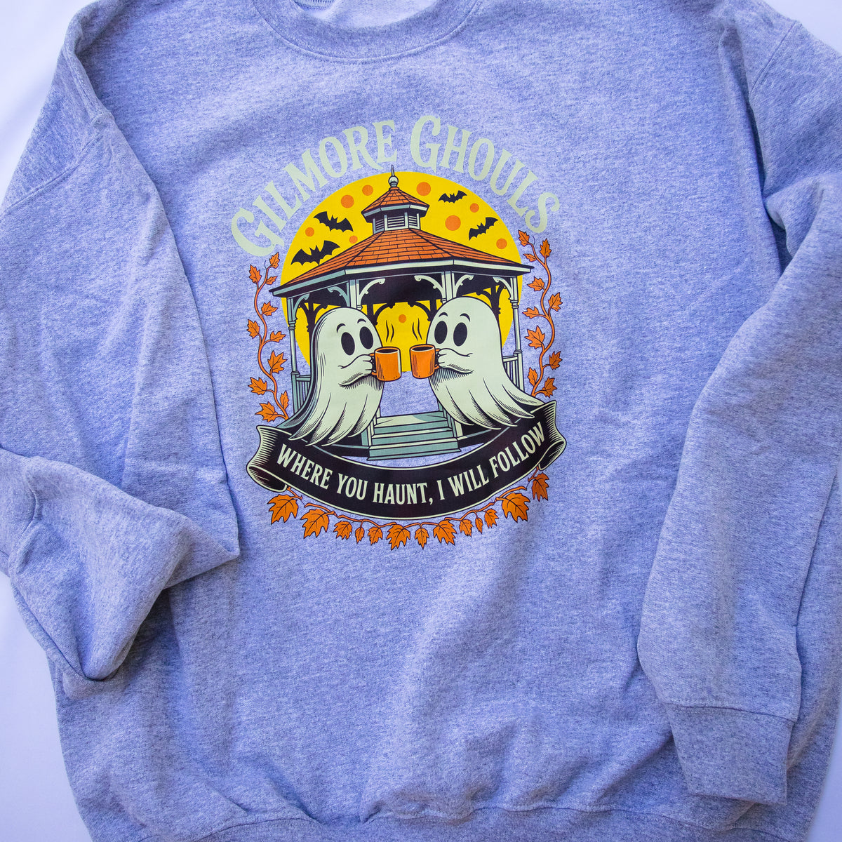 A heather gray crewneck sweatshirt featuring a colorful graphic print of two cartoon ghosts at a gazebo with the text GILMORE GHOSTS WHERE YOU HAUNT, I WILL FOLLOW'.