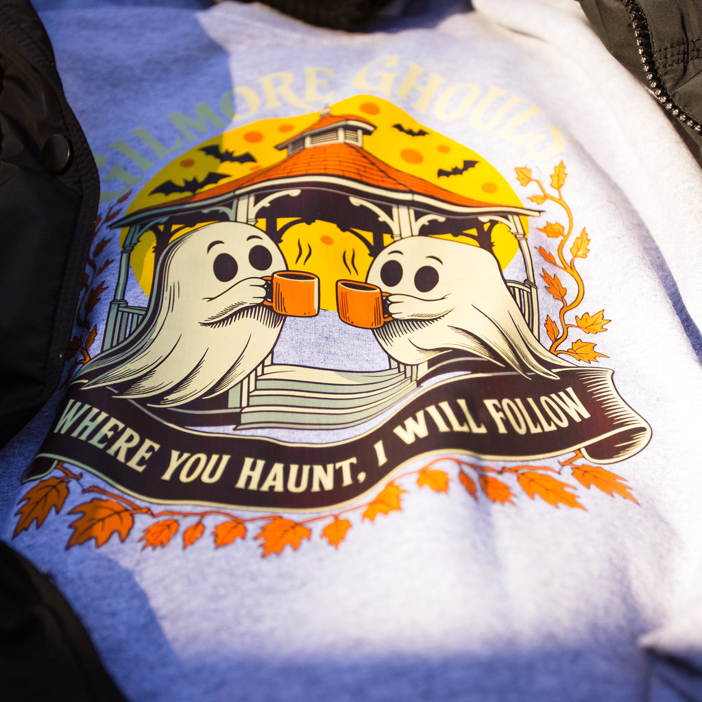 A close-up of a light blue heather t-shirt featuring a Halloween graphic of two ghosts holding orange mugs in front of a haunted house, with the text 'WHERE YOU HAUNT, I WILL FOLLOW'.