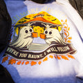 A close-up of a light blue heather t-shirt featuring a Halloween graphic of two ghosts holding orange mugs in front of a haunted house, with the text 'WHERE YOU HAUNT, I WILL FOLLOW'.