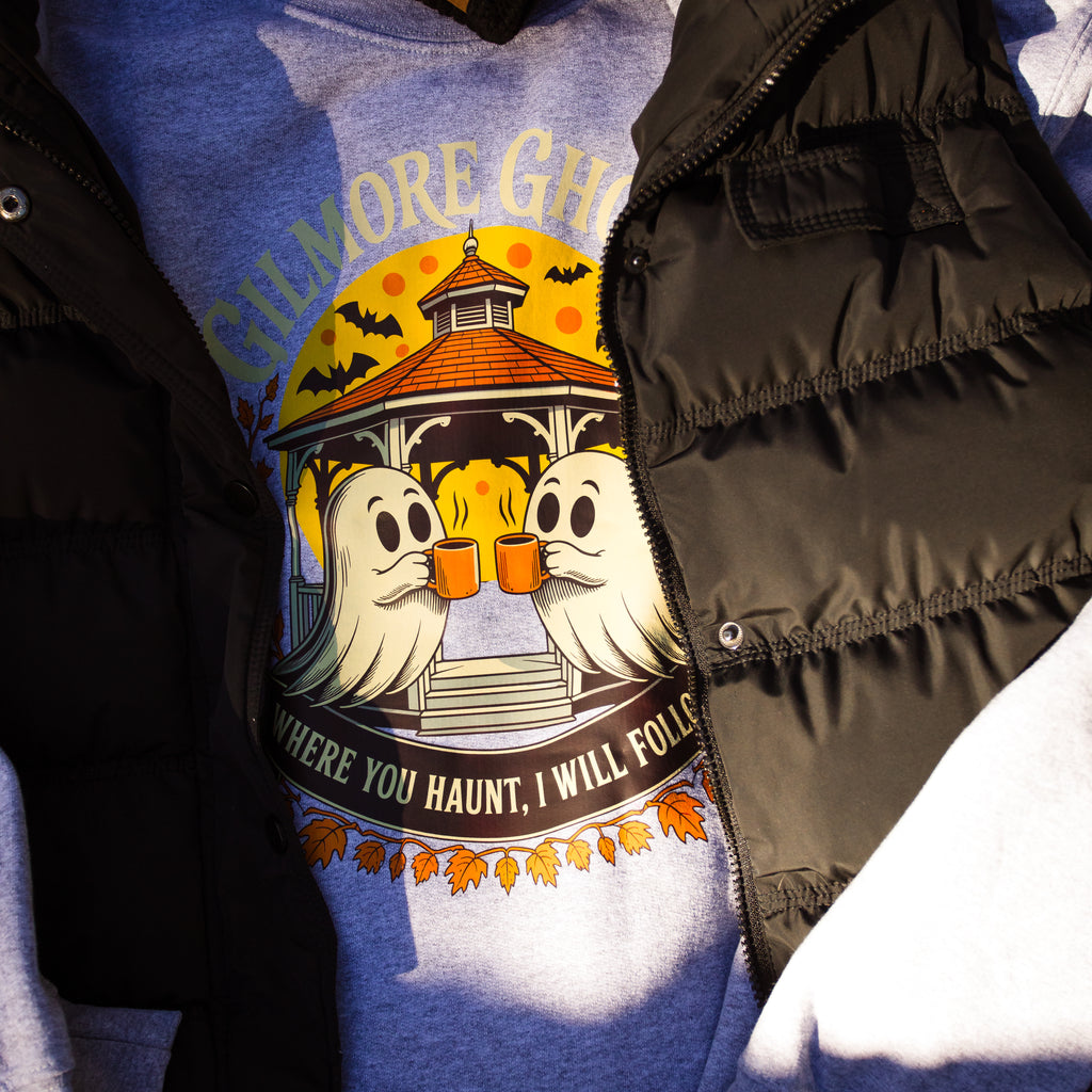 A close-up of a heather gray graphic t-shirt featuring a Halloween design with two ghosts holding orange mugs in front of a gazebo, styled under a black puffer jacket.