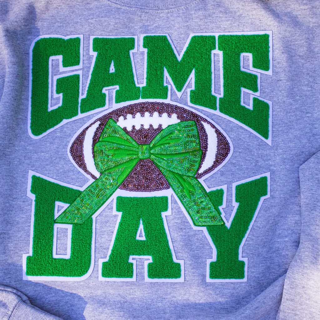 A close-up of a heather gray sweatshirt featuring a green and white 'GAME DAY' graphic with a football and a glittery green bow in the center.