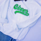 A white crewneck sweatshirt featuring a green sequined 'Cheer' script across the chest, styled flat with blue jeans on a light blue background.