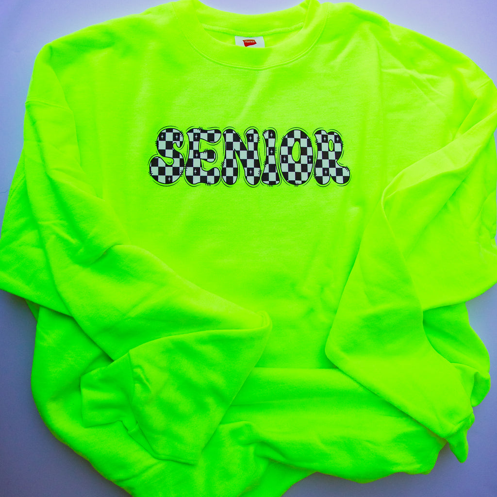 A neon green long-sleeve t-shirt with 'SENIOR' printed across the chest in a black and white checkered font, laid flat on a gray background.
