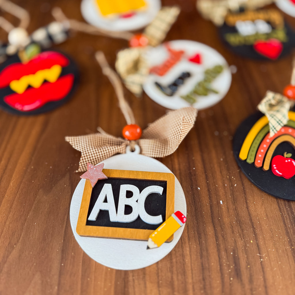 A close-up of a round white ornament featuring a black chalkboard with white letters 'ABC' and a yellow pencil, tied with a beige ribbon and orange bead, displayed on a wooden surface with other colorful ornaments blurred in the background.