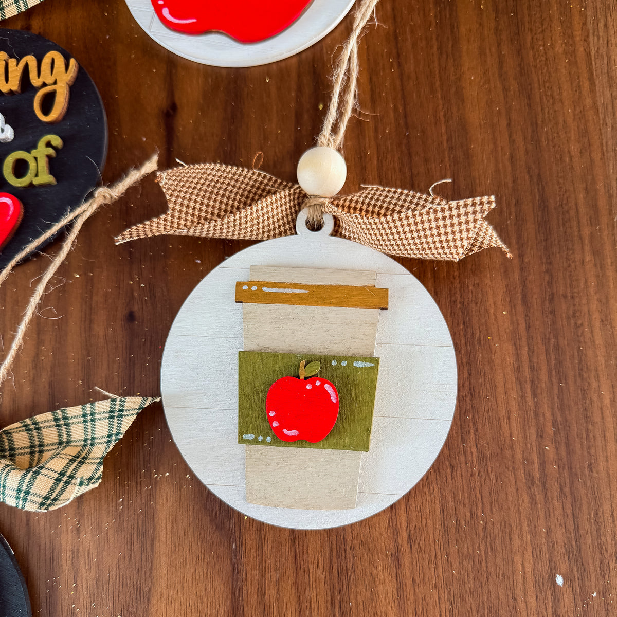 A circular wooden ornament decorated to look like a takeaway coffee cup with a red apple on the front, hanging on a wooden surface with a brown gingham ribbon and jute string.