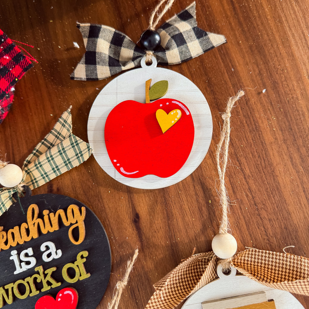A close-up of a decorative autumn-themed ornament featuring a red apple with a yellow heart-shaped accent on a white round background, hanging from a black-and-cream plaid ribbon on a wooden surface.