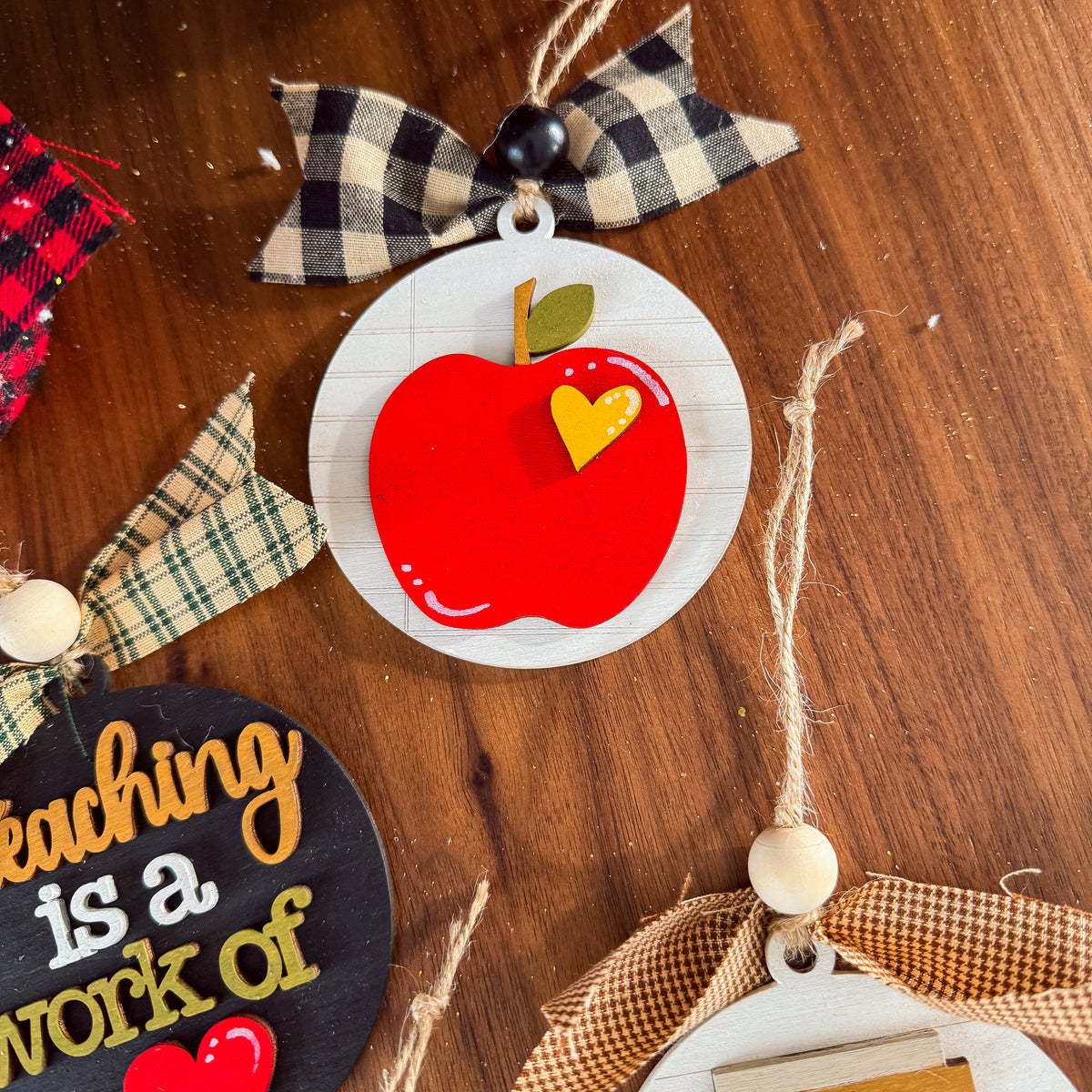 A close-up of a decorative autumn-themed ornament featuring a red apple with a yellow heart-shaped accent on a white round background, hanging from a black-and-cream plaid ribbon on a wooden surface.