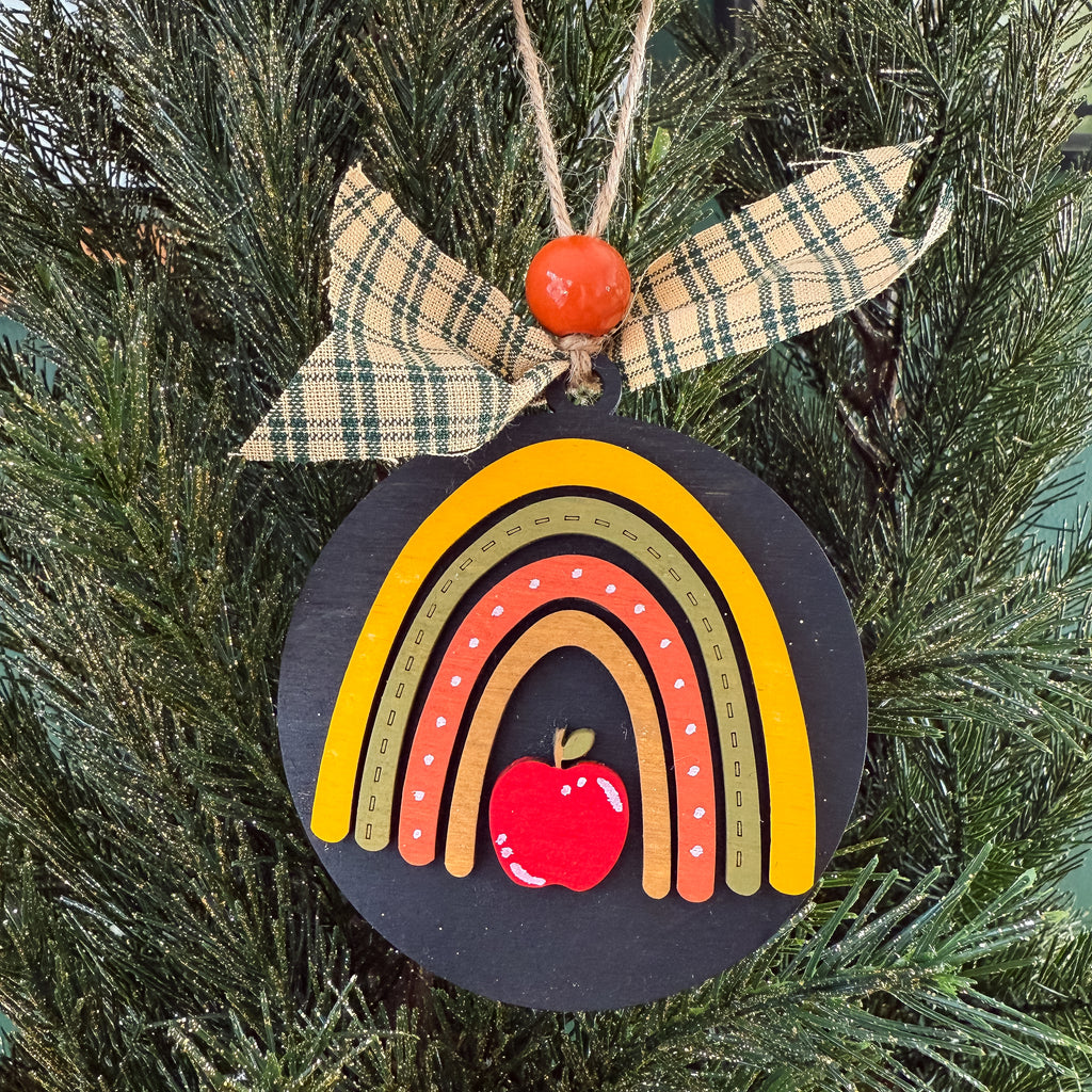 A round black wooden Christmas ornament featuring a colorful rainbow design with a red apple at the center, hanging from a plaid ribbon bow on a green Christmas tree.