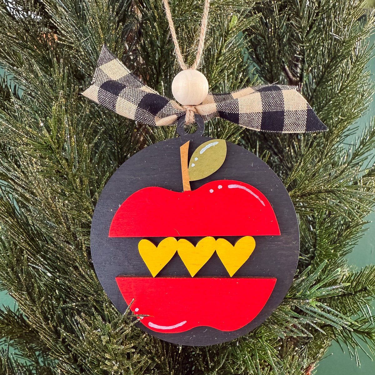 A circular black wooden apple ornament hanging on a green Christmas tree. The ornament is designed like a red apple with yellow heart-shaped teeth and a small green leaf, tied with a black-and-beige plaid ribbon and a wooden bead at the top.
