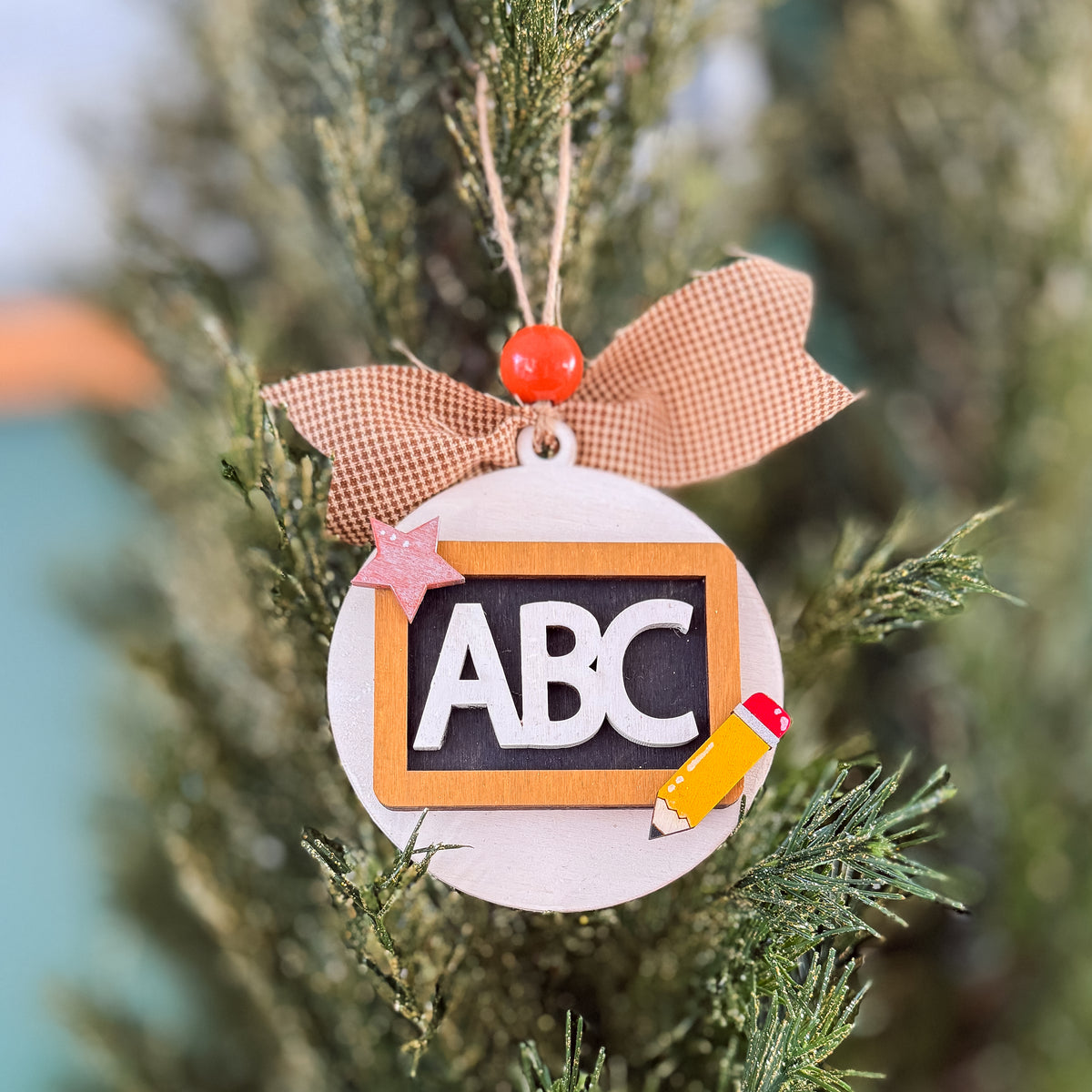 A round Christmas ornament shaped like a chalkboard with the letters ABC in white, hanging on a tree branch. The ornament has a tan ribbon bow with a small orange bead, a pink star, and a yellow pencil detail.