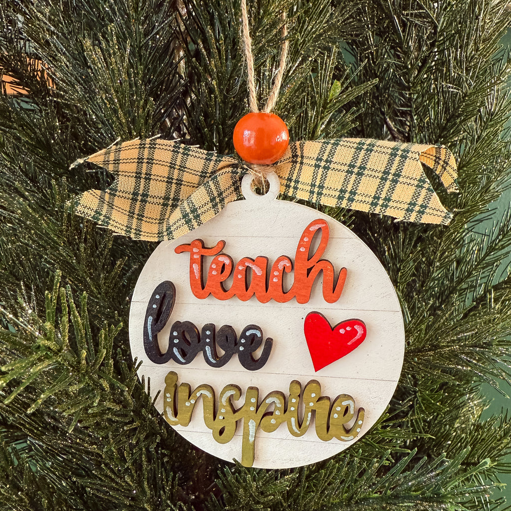 A round white wooden Christmas ornament hanging on a green pine tree, decorated with a yellow plaid ribbon bow, an orange bead, and raised lettering that reads 'teach love inspire' with a red heart.