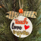 A round white wooden Christmas ornament hanging on a green pine tree, decorated with a yellow plaid ribbon bow, an orange bead, and raised lettering that reads 'teach love inspire' with a red heart.