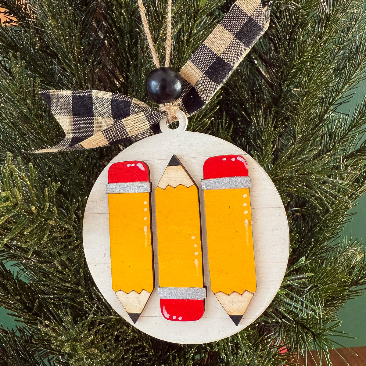 A round wooden Christmas ornament shaped like a stack of three yellow pencils with red erasers, hanging on a green Christmas tree with a black-and-beige plaid ribbon.