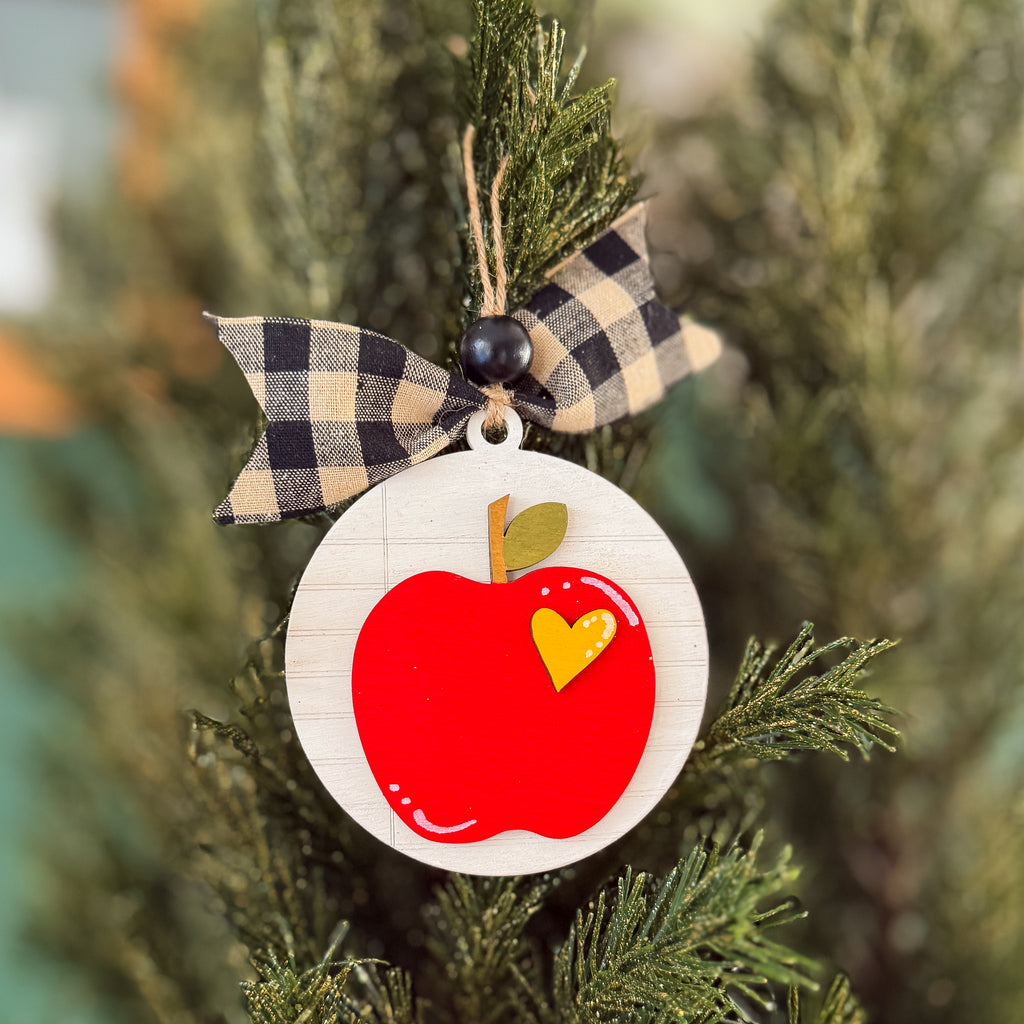 A round wooden apple ornament hanging on a pine tree. The ornament has a white wood-grain background, a red apple with a small yellow heart-shaped accent, and is tied with a beige and black buffalo plaid ribbon.