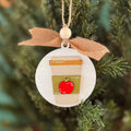 A round wooden Christmas ornament shaped like a takeaway coffee cup with a red apple on the sleeve, hanging from a tan ribbon on a blurred evergreen tree.