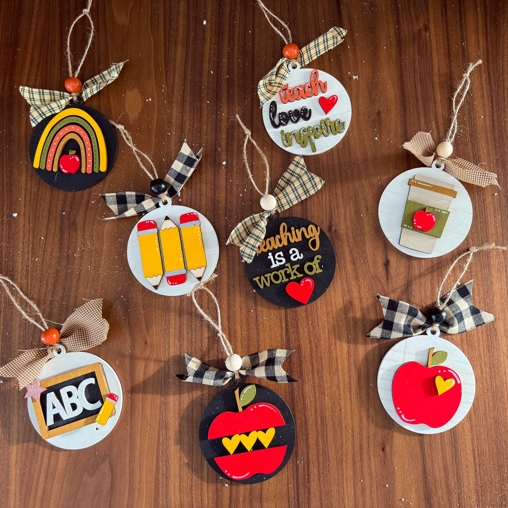 A set of nine school-themed paper ornaments arranged on a dark wood background. The ornaments feature various designs including a rainbow, three yellow pencils, an ABC sign, a red apple, a coffee cup, and text messages like 'teach love together' and 'teaching is a work of art'.
