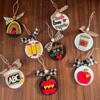 A set of nine school-themed paper ornaments arranged on a dark wood background. The ornaments feature various designs including a rainbow, three yellow pencils, an ABC sign, a red apple, a coffee cup, and text messages like 'teach love together' and 'teaching is a work of art'.