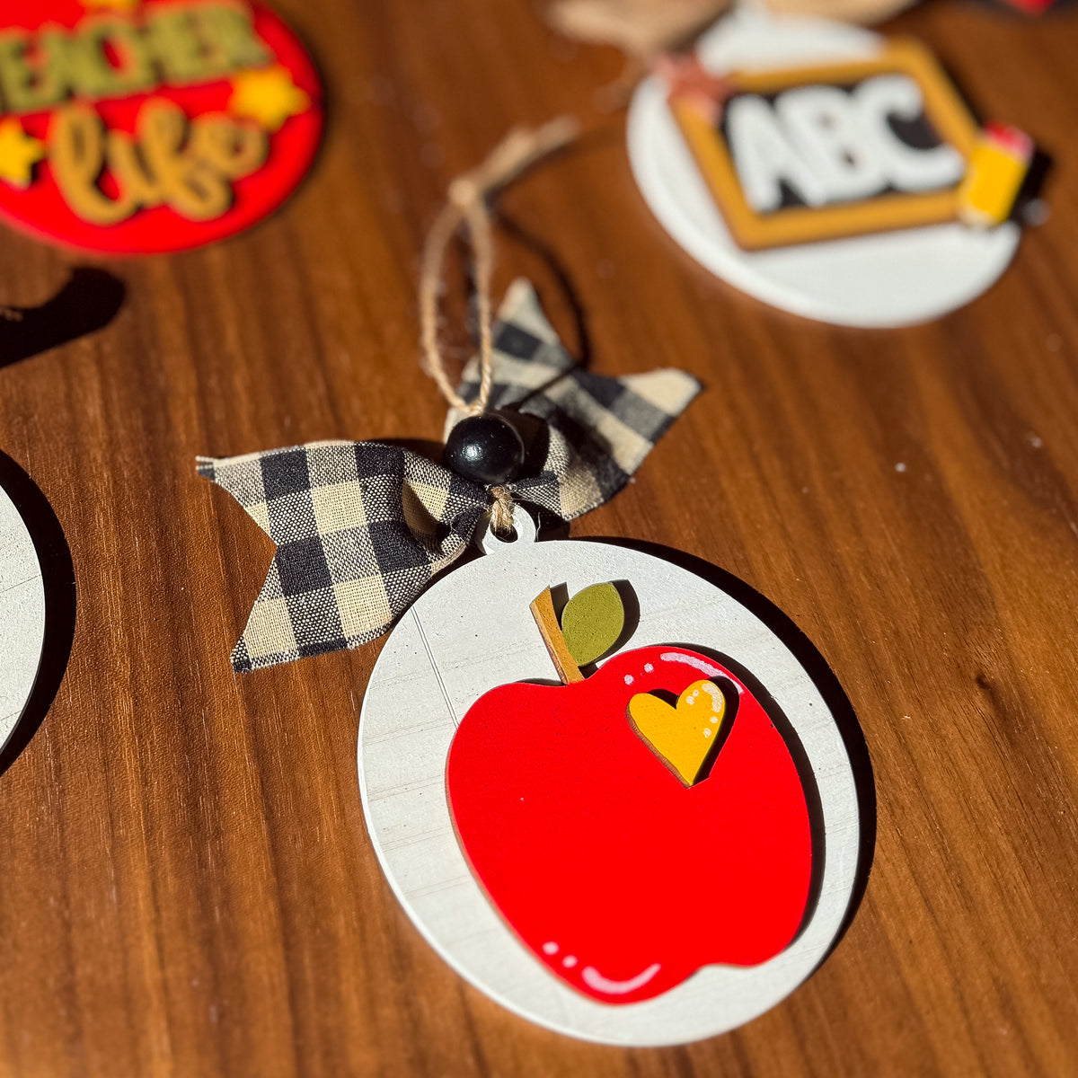 A close-up of a round wooden apple ornament with a red apple design, small yellow heart accent, and a black-and-beige gingham bow on a twine string, resting on a wooden surface.